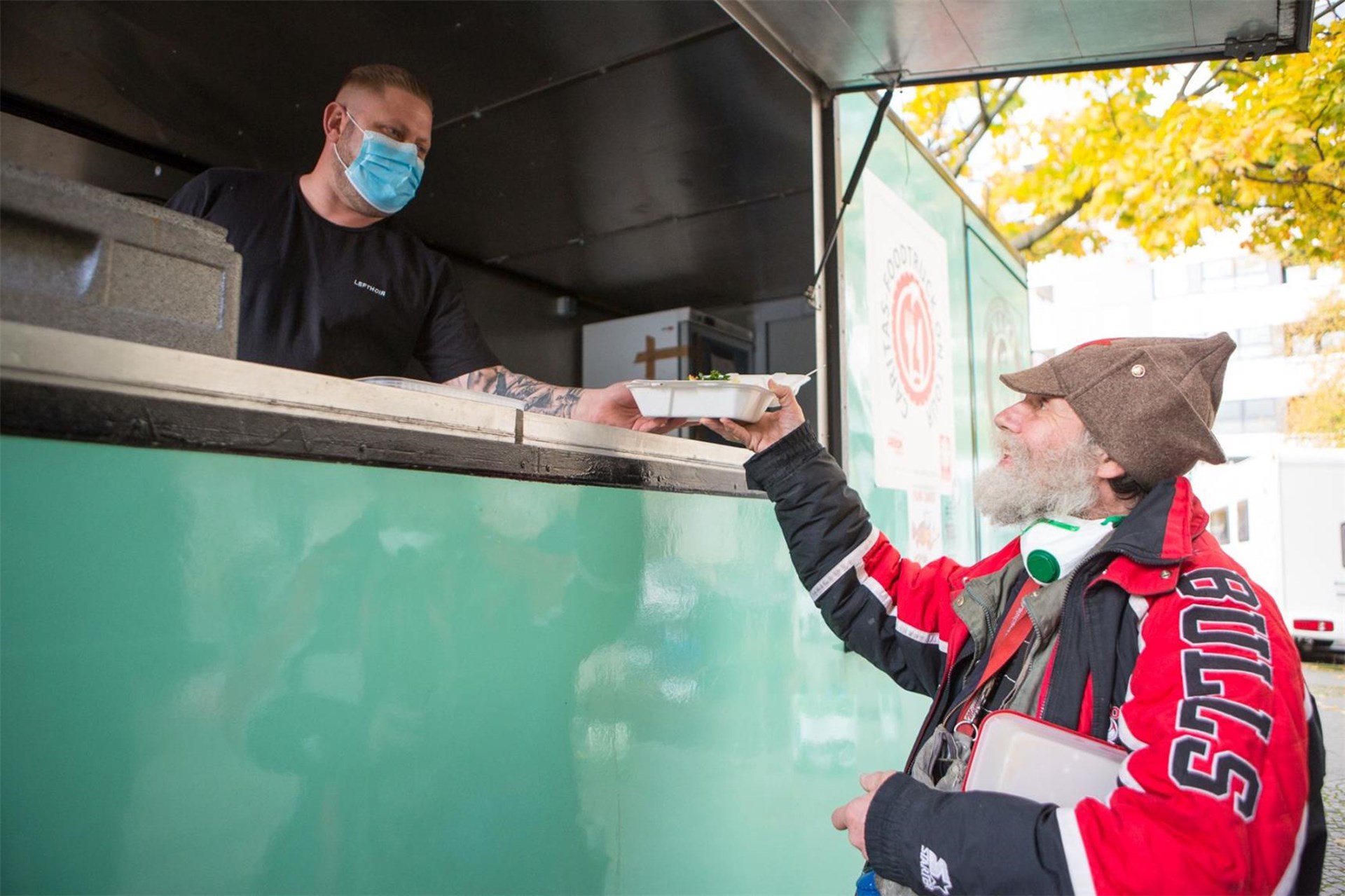 Mann mit Maske reicht Essen in Pappschale an älteren Mann mit roter Jacke und Mütze an einem Essensstand.